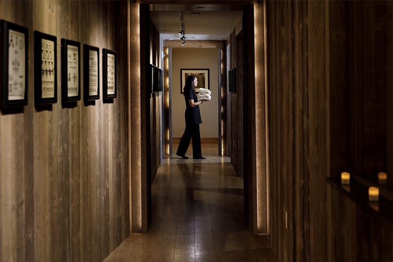 A spa worker wearing all black carrying fluffy white towels down a wooden paneled hallway.