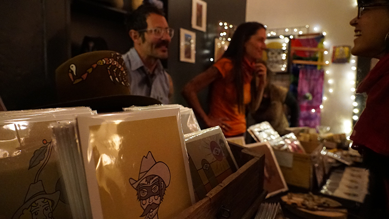 Local artist greeting cards sit in front of vendors in a dimly lit room with Christmas lights