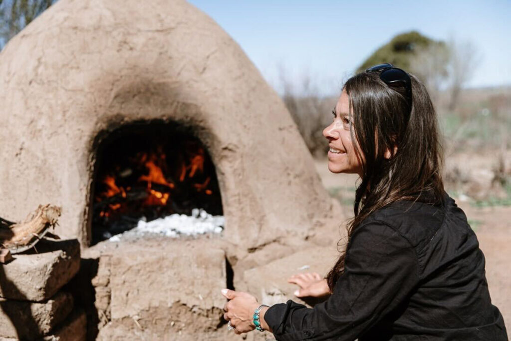 A woman sites in front of an horno, an adobe outdoor oven with a wood fire inside