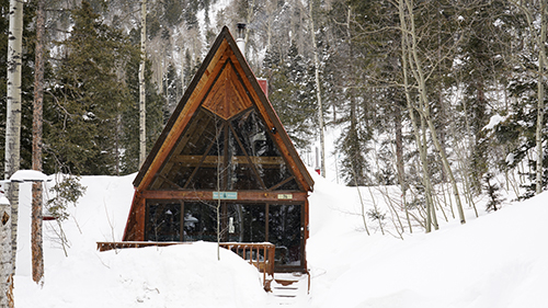 A wooden A-frame cabin surrounded by tall fir trees and a thick layer of snow.