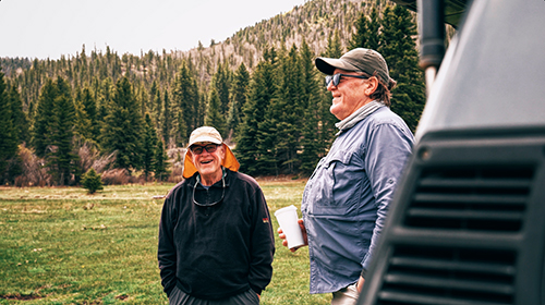 Two Taos fly fishing guides stand in a field in the mountains talking to each other.