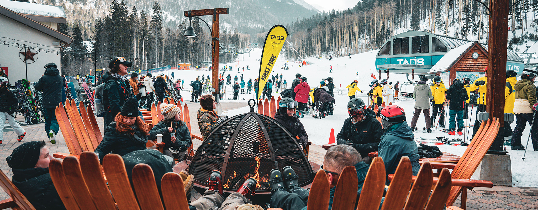 A group of skiers take a break in Adirondack chairs around the fire pit while watching skiers on the mountain.