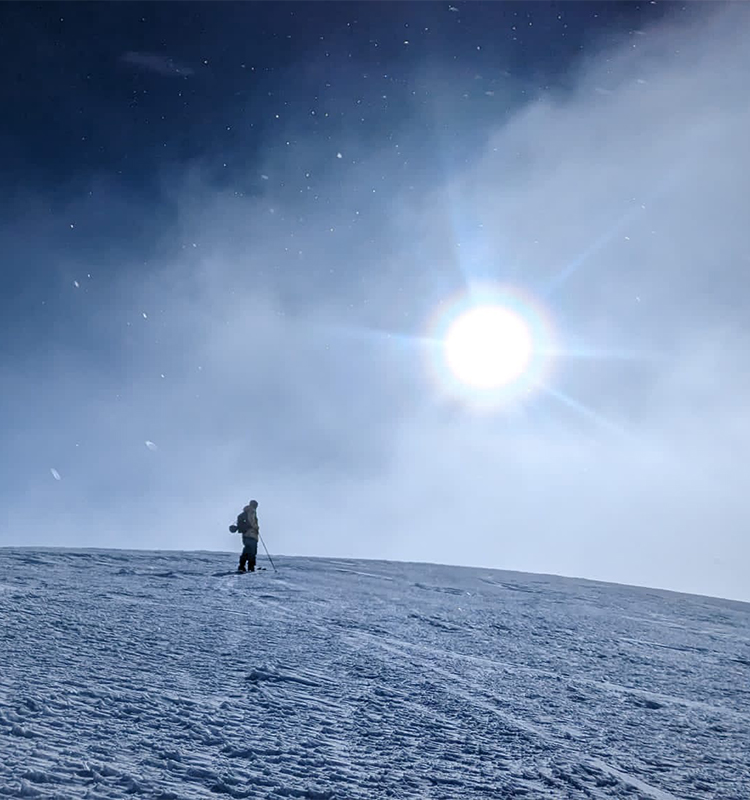 A lone skier stands on a barren mountain top with cold smoke blowing in the morning sun.
