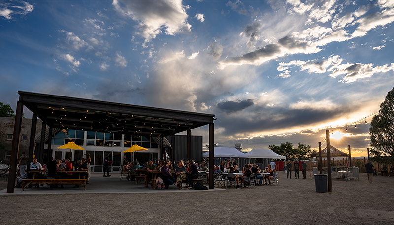 An expansive sky of wispy clouds and warm glow of sunset behind an industrial style outdoor patio filled with bar patrons.