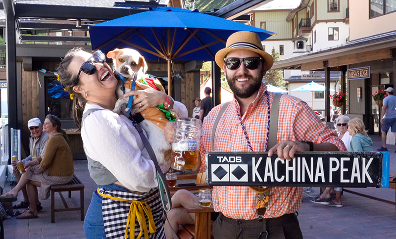 A man and woman in traditional Bavarian dress laugh while holding a dog and a "Kachina Peak" sign.