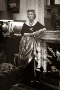 A black and white photos of a stylish woman leaning against a fireplace mantle.