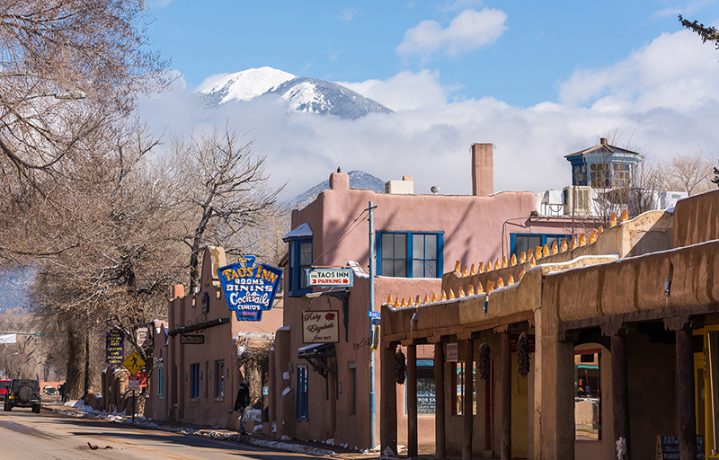 A street of old style adobe buildings sit in front of a snow covered mountain with low hanging clouds.