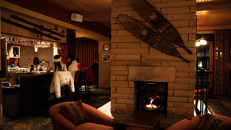 A dimly lit bar and lounge with a fireplace in the forefront and plush chairs.