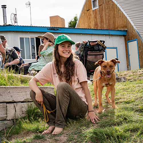 A woman sits barefoot on a small grassy hill smiling next to her dog.