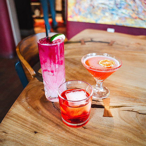 Three colorful cocktails in different glasses sit on a live edge wood table.