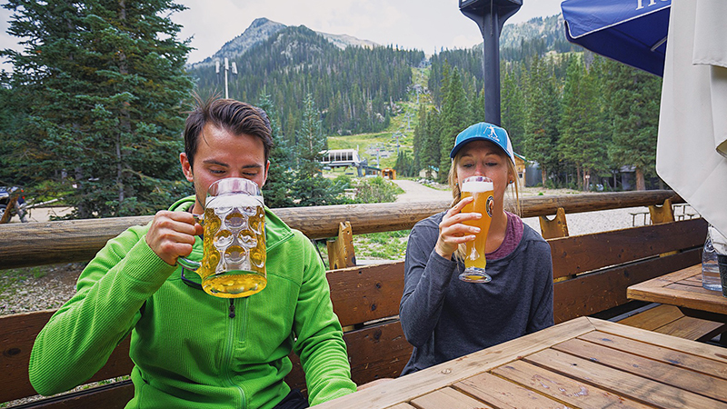 A man and woman sit on an outdoor deck drinking large German beers in front of a mountain backdrop.