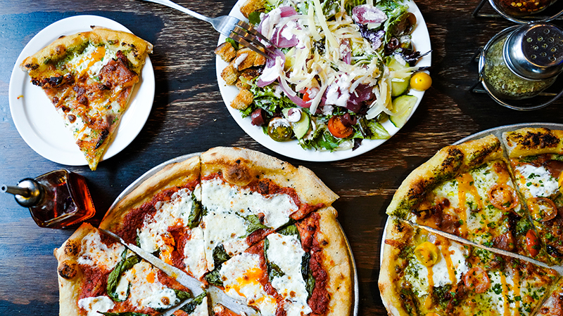 Wood fired pizzas and extravagant salads sit on a table.