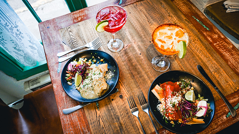 An overhead view of two black dinner plates on a wooden table with tamales and colorful cocktails on the side.