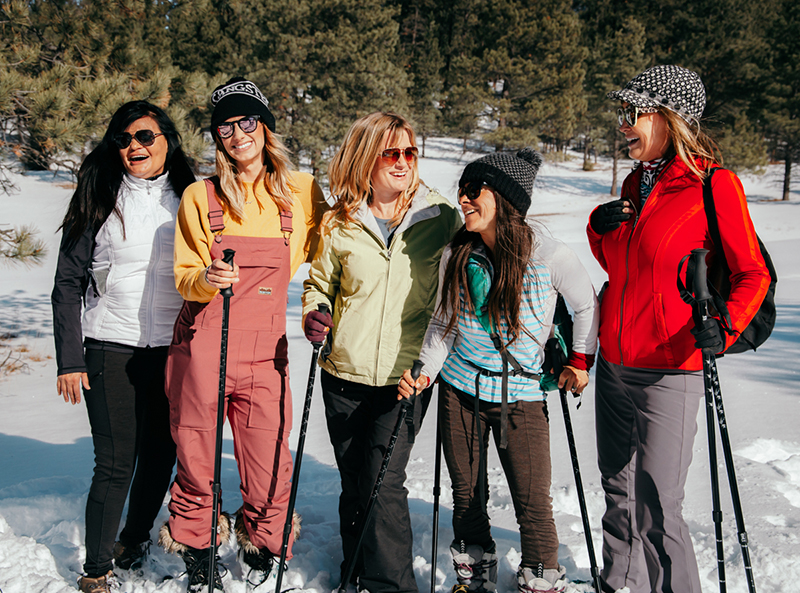 Five women laugh together in a snowy field wearing snowshoes.
