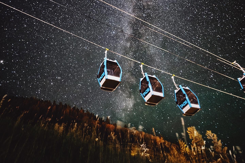 Three ski gondolas hang from a cable at night with the Milky Way shining behind them.