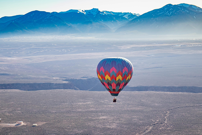 A colorful hot air balloon floats high above the ground with snow capped mountains in the distance.