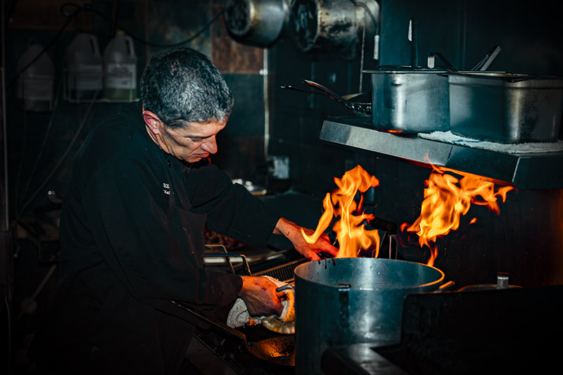 A concentrated chef wearing all black works at a flaming stove surrounded by large pots.