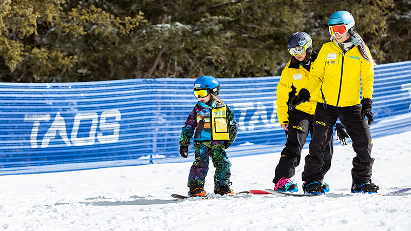A young child on a snowboard is followed by two instructors in yellow jackets down a slope.