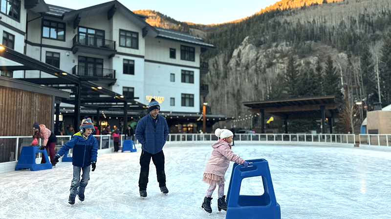 A father and two children ice skate, with one child using an assist.