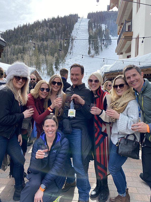 A group of smiling friends, one wearing a fur hat, smile with a bottle of wine in front of a snowy mountain.