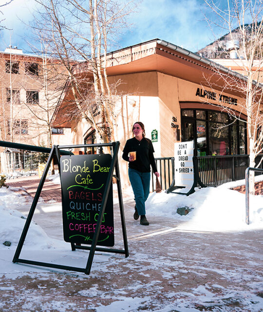 Woman walking with orange juice and bagel in Taos Ski Valley.