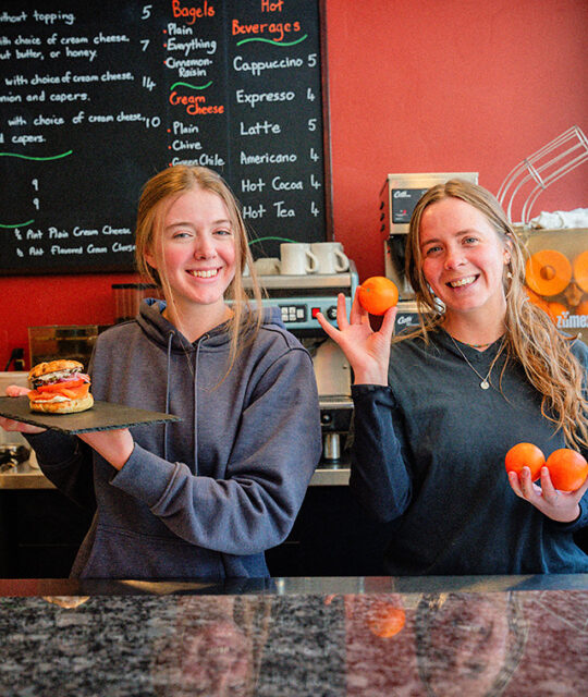 Happy servers holding a bagel sandwich and oranges at The Blonde Bear Cafe.