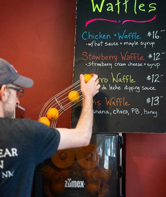 Blonde Bear Cafe Waffle menu on chalkboard with man adding oranges to the fresh squeezed orange juice machine.