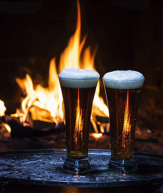 Two pilsner glasses in front of a fireplace.