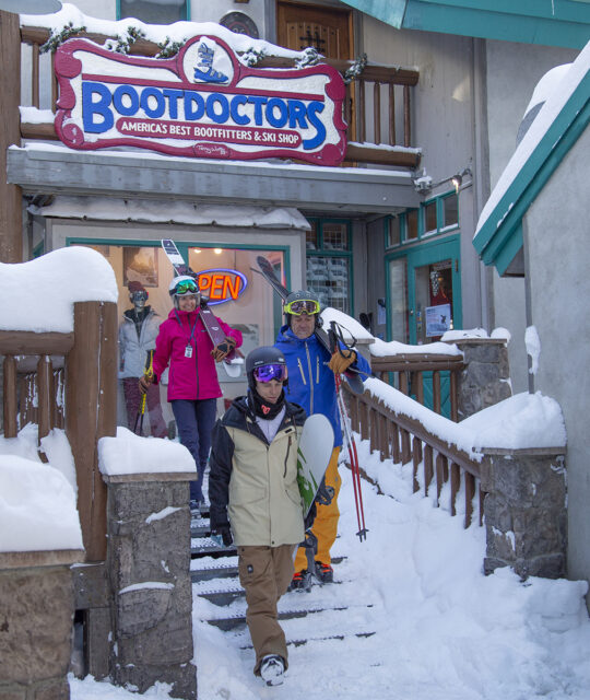 Skiers walk past BootDoctors store with their gear in the snow.