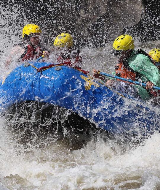 Rafters in whitewater rapids with yellow helmets in a blue raft.