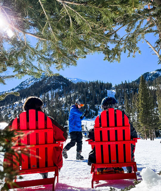 Skiers sitting in red Adirondack chairs looking up at ski mountain on a sunny day.