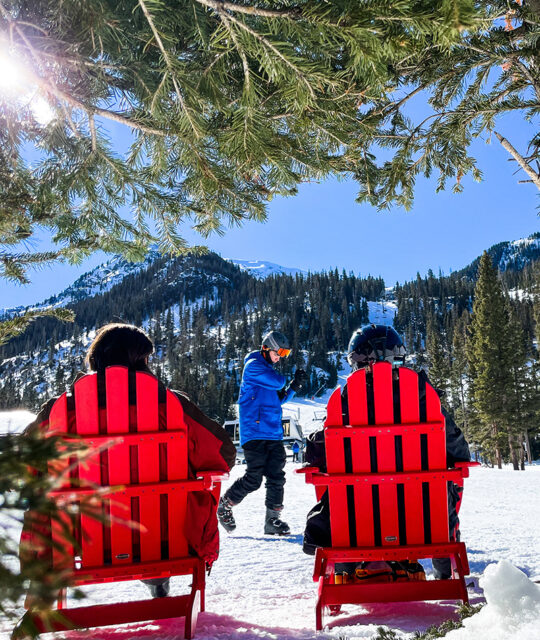 Skiers sitting in red Adirondack chairs looking up at ski mountain on a sunny day.