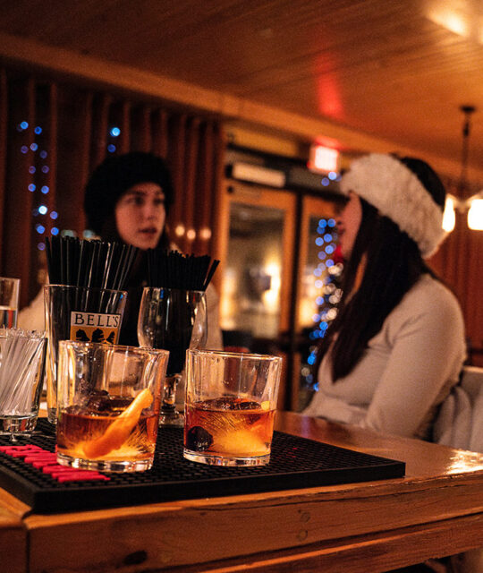 Two women in winter ski clothes seated at the bar at The Blonde Bear Tavern with drinks in the foreground..