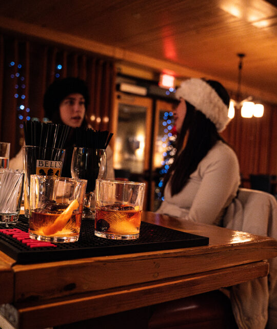 Two women in winter clothes seated at a bar with drinks.