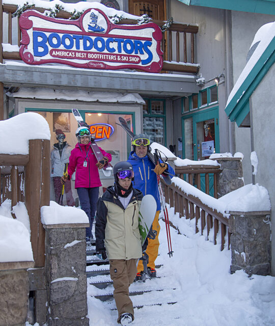 Skiers walk past Bootdoctors store with their gear in the snow.