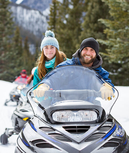 Couple on snowmobile tour with mountain background.
