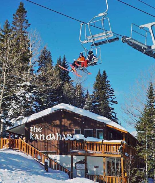 Sunny bluebird day with skiers on lift and the snowy Kandahar Condominiums behind.