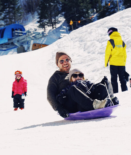 A father and child ride a sled together at Taos sledding hill.