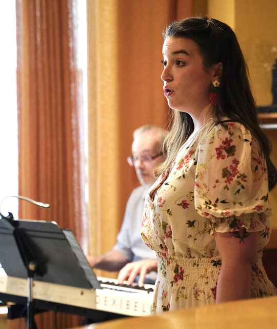 Young female opera singer performing with a pianist in the background.