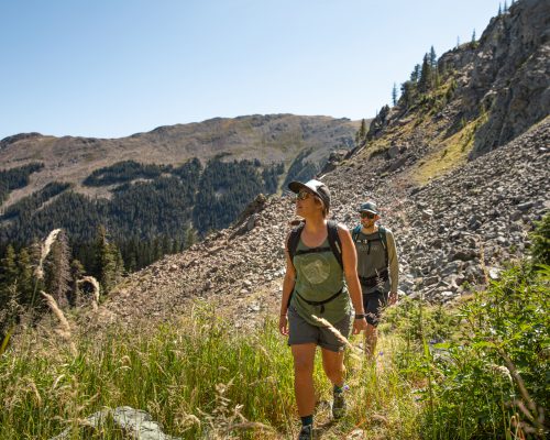 A woman and man hike through tall grass on a rocky mountainside