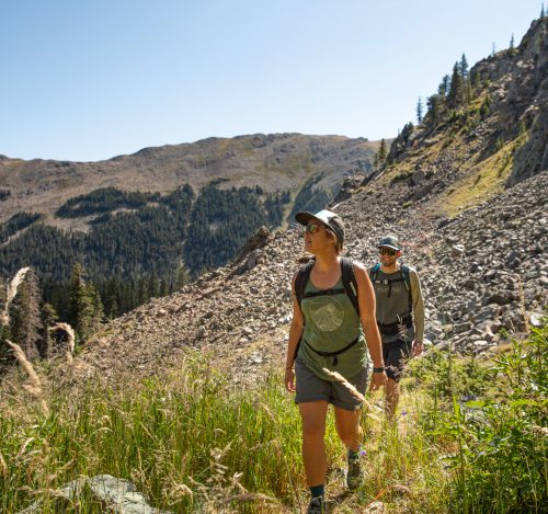 A woman and man hike through tall grass on a rocky mountainside