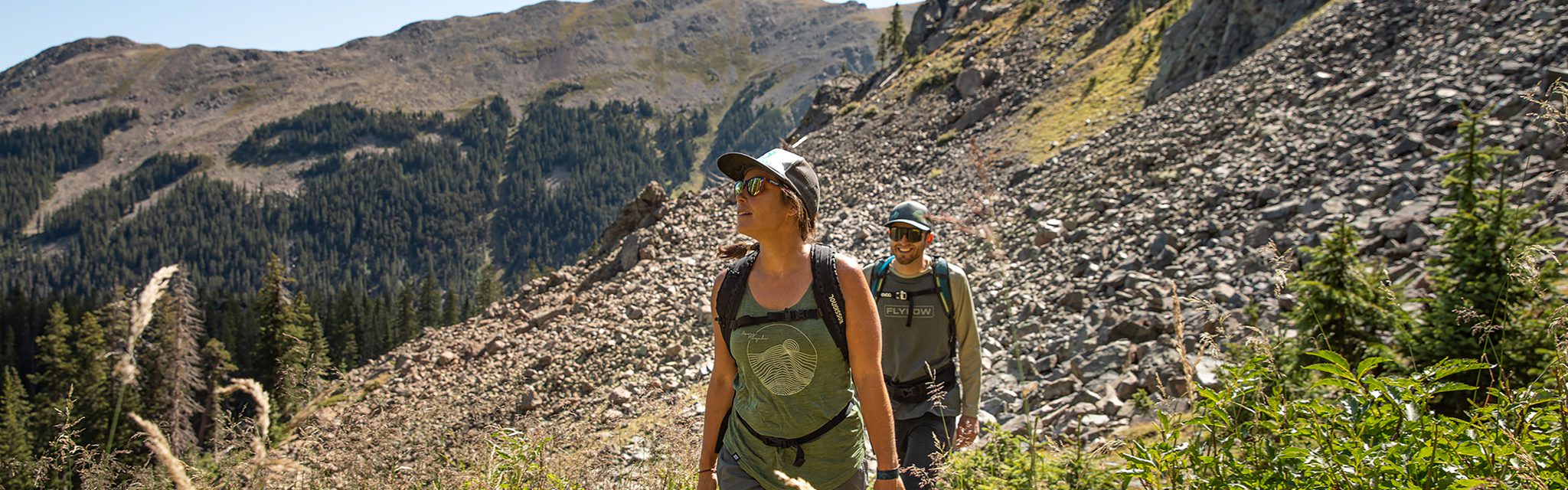 A man and woman hike in front of a rocky mountain face in Taos Ski Valley