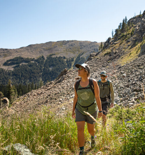 A man and woman hike in summer in front of a rocky mountain face in Taos Ski Valley