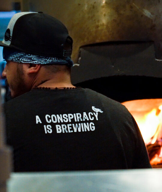 Cook at wood fired oven wearing a Conspiracy is Brewing t-shirt.