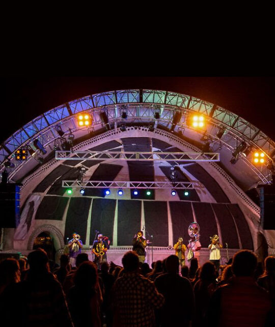 An arched outdoor concert stage, lit up in front of a packed crowd.