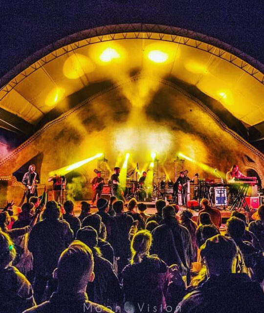An arched outdoor concert stage, lit up yellow in front of a packed crowd