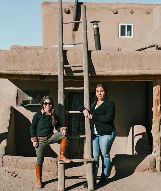 Two women guides for Taos Pueblo tours posing with a traditional ladder.
