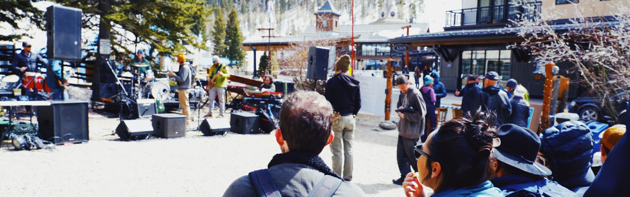 A crowd of people eat and watch a band playing an outdoor show in winter, with ski slopes behind them.
