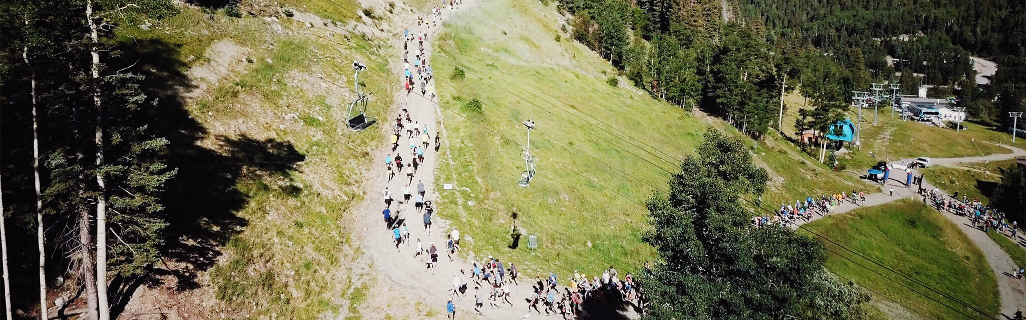 An overhead view of a trail run starting line, with runners climbing a steep trail under a chairlift in summer.
