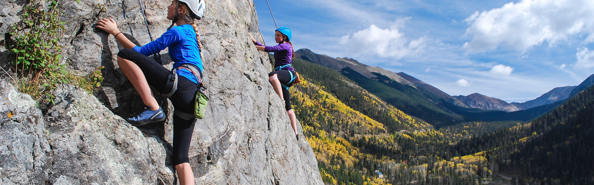 Two teenage girls climb a rock face above a yellow aspen meadow.